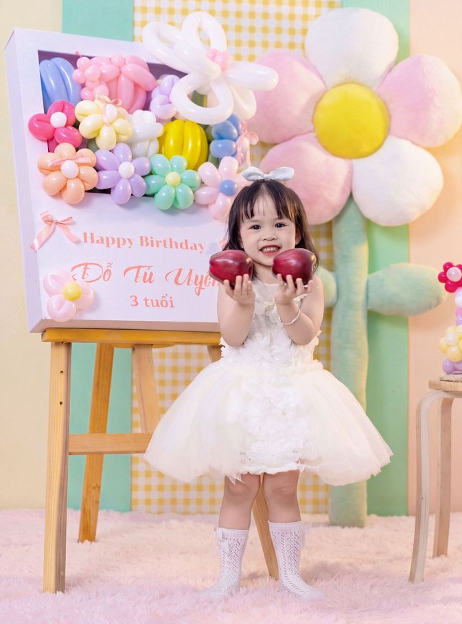 Child in a white dress holding apples at a pastel balloon-decorated birthday setup