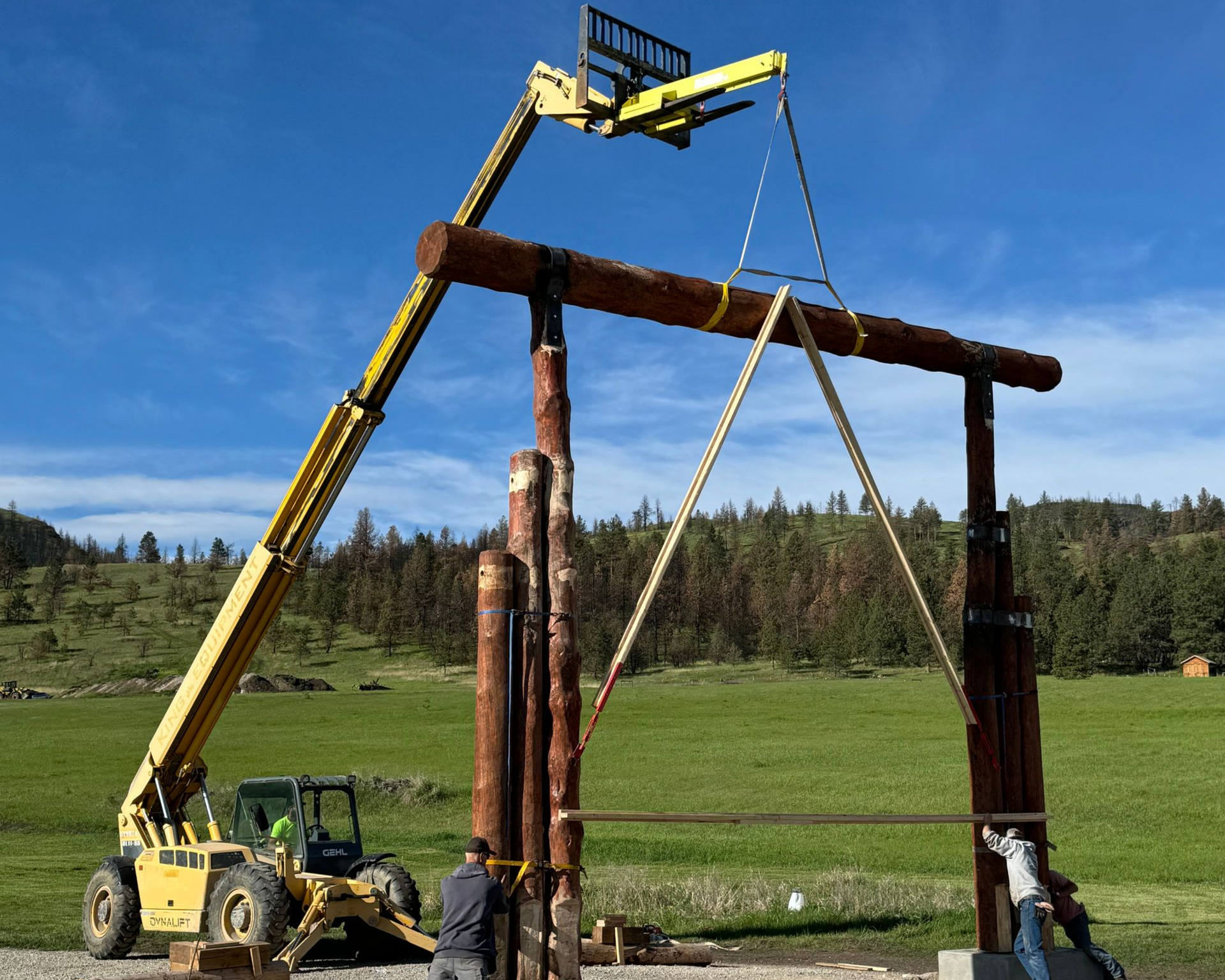 Forklift lifting a large wooden beam for a gate. Construction workers stand nearby outdoors with a green field and blue sky.