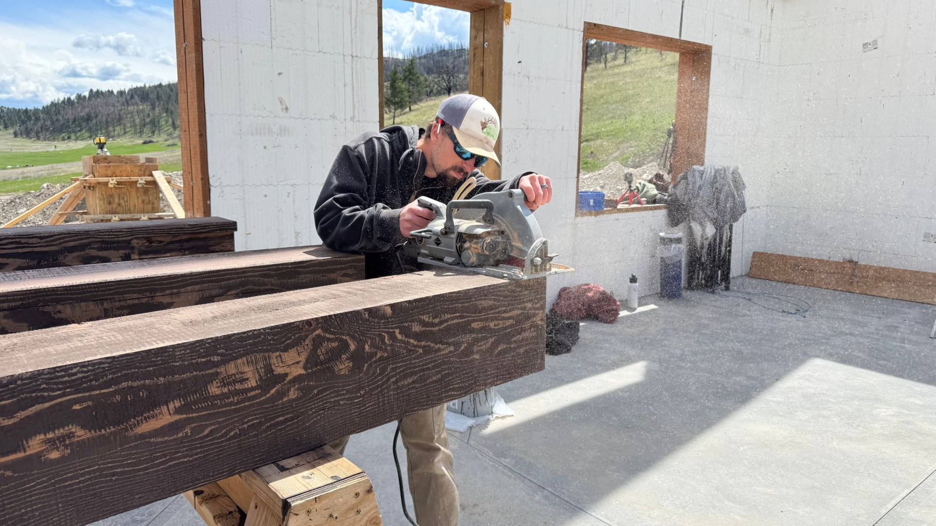 Man cuts wooden beam with a saw outside a building under construction.