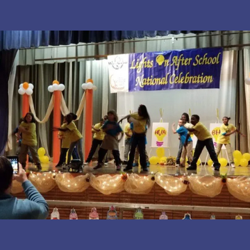 A group of children are dancing on a stage in front of a sign that says lights on after school national celebration