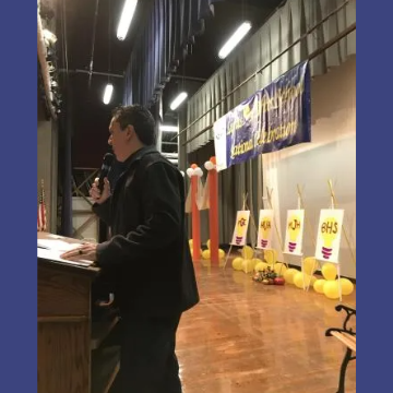 A man stands at a podium with a microphone in front of a banner that says hhs