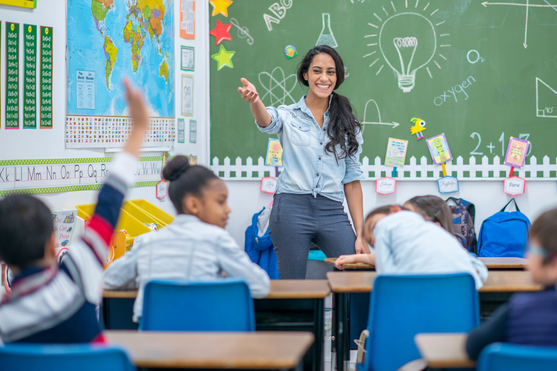 A teacher is standing in front of a classroom of children raising their hands to answer a question.