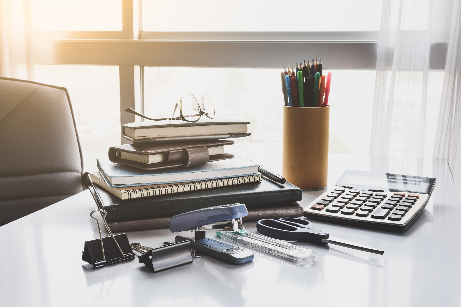 A desk with a stack of books , a calculator , a pen holder and a pencil holder.