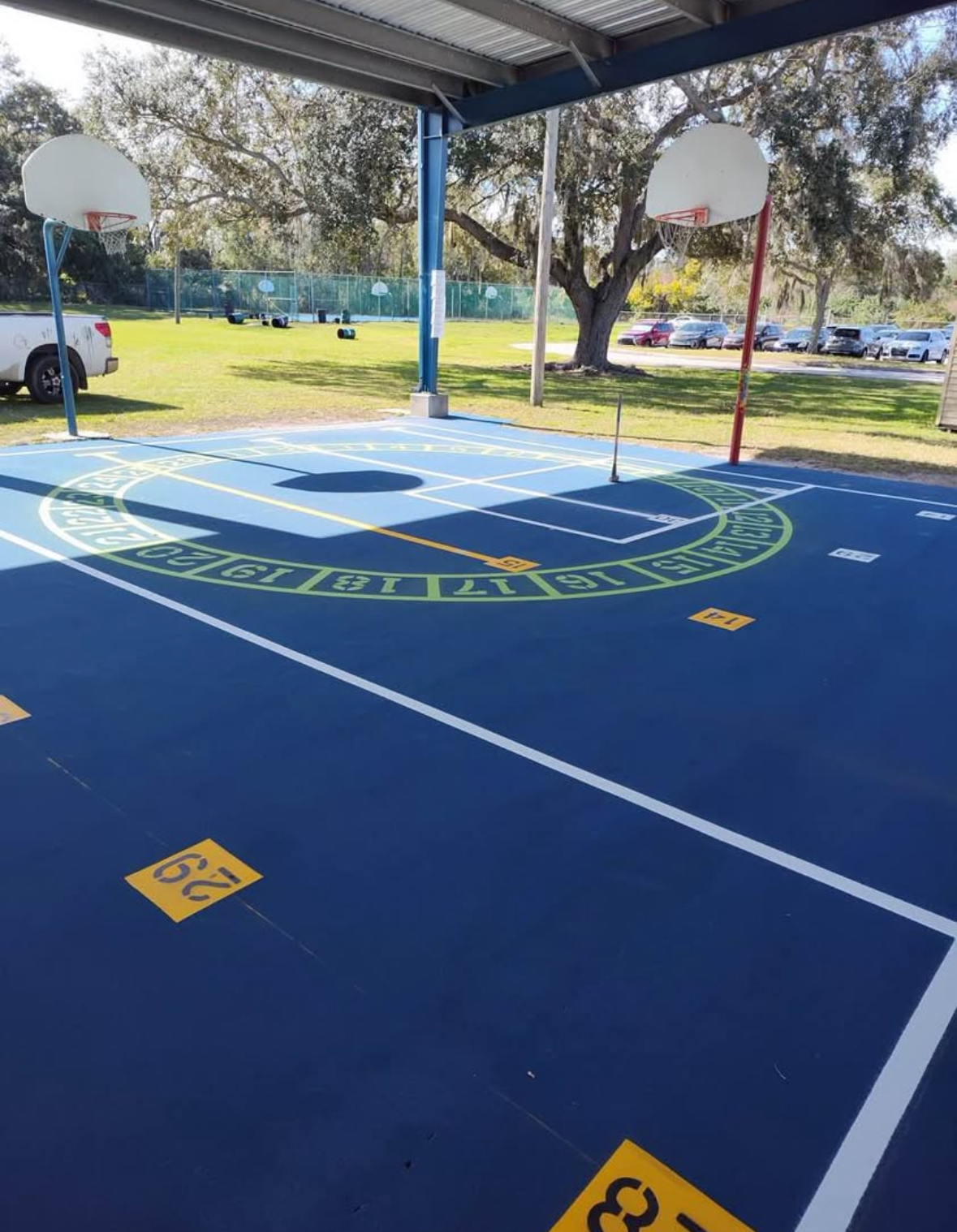 Covered outdoor basketball court with blue flooring, white hoops, and trees in the background