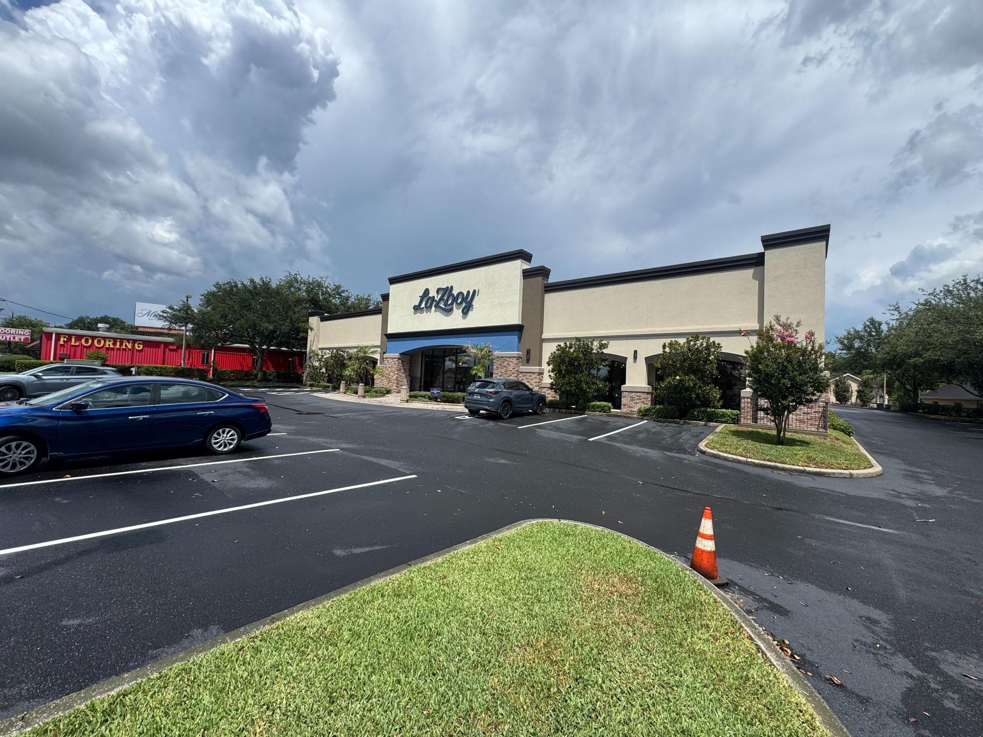 Strip mall storefront with beige facade, parking lot, parked cars, and a cloudy sky