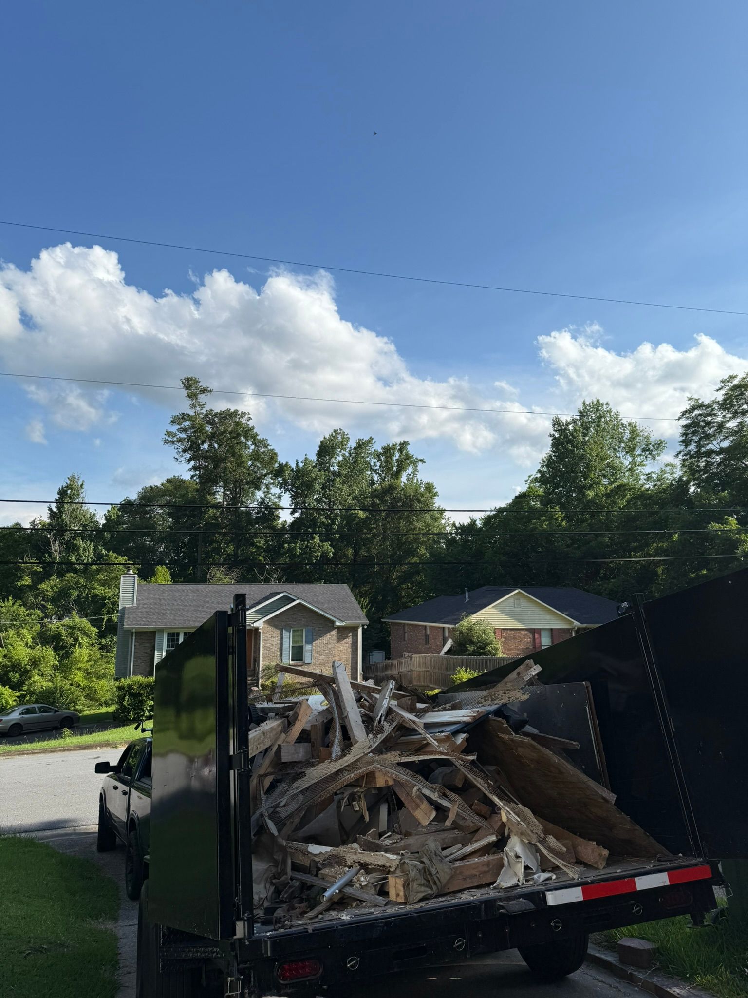 Truck bed filled with debris parked on a street in front of houses with a blue sky and clouds above.