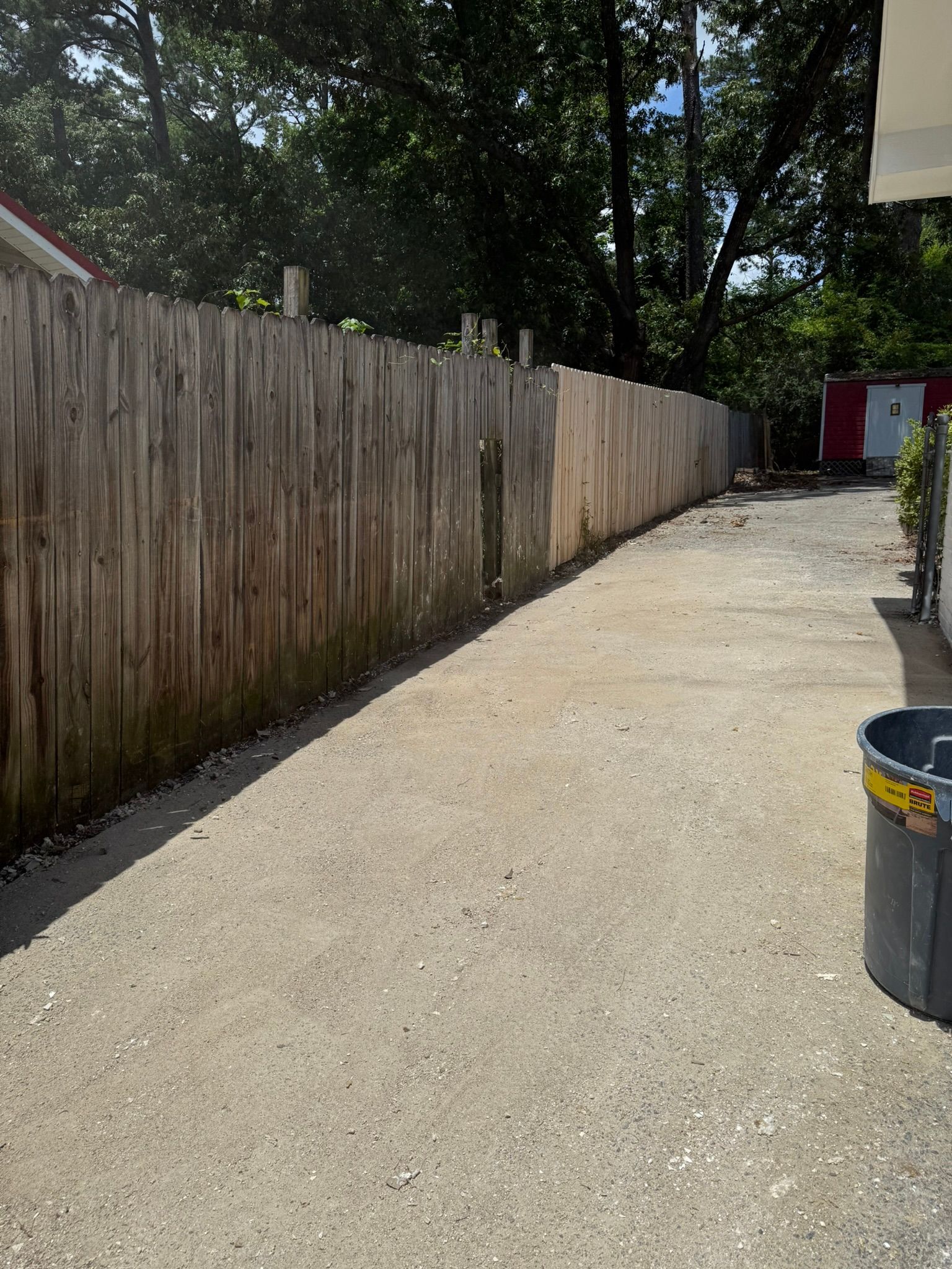 Gravel driveway alongside a weathered wooden fence, leading towards trees and a red building.