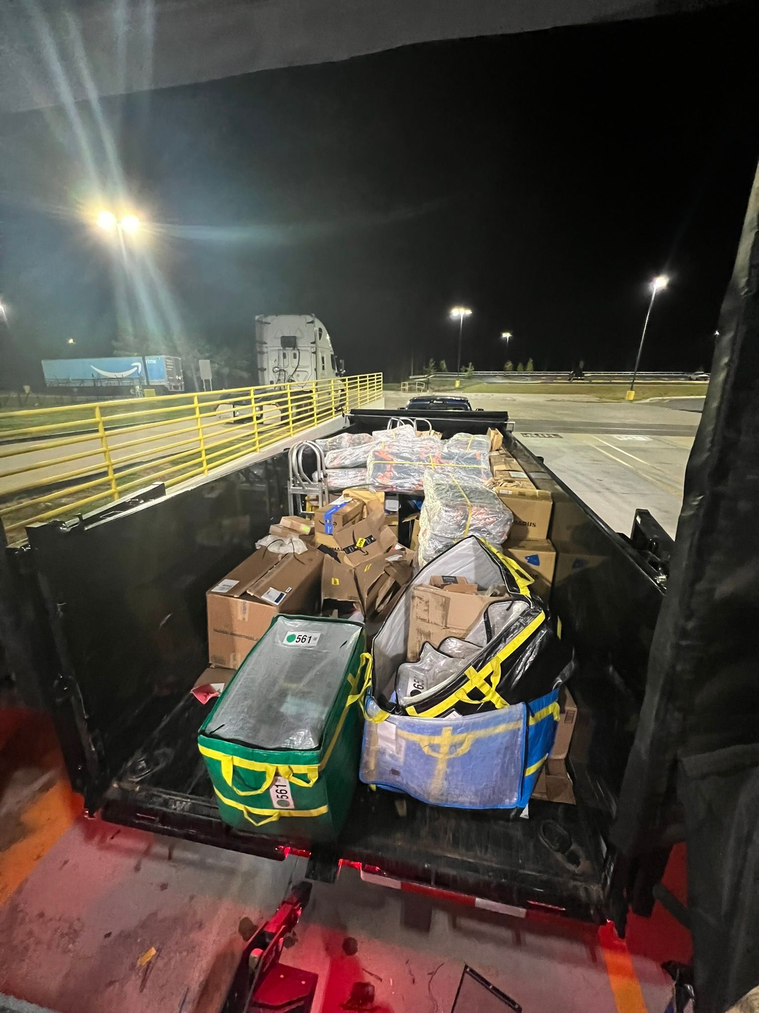 Cargo trailer filled with boxes and bags, likely packages. Nighttime, exterior setting with lights.