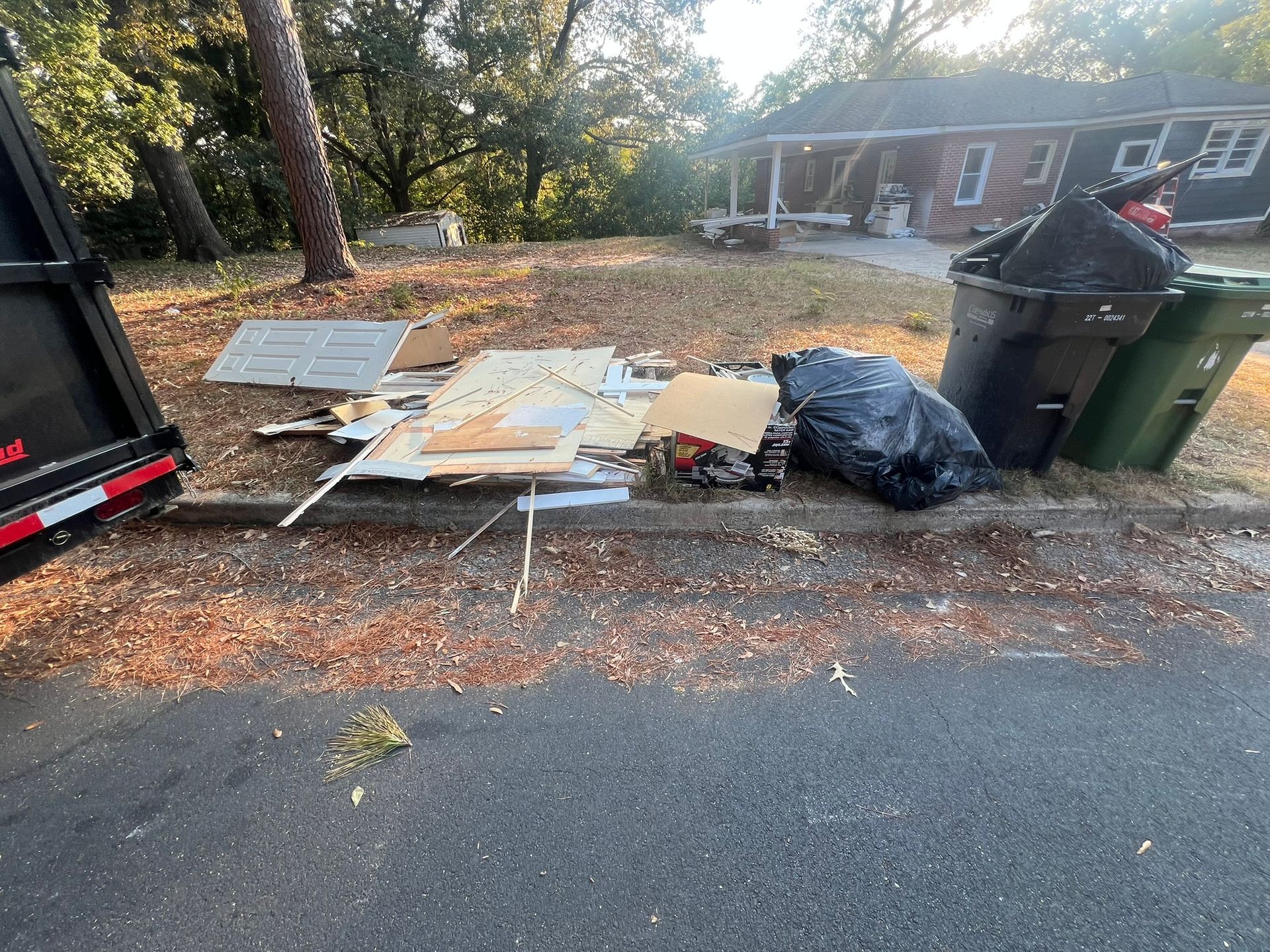 Debris pile on curb with dumpster, trash cans, and house in background.