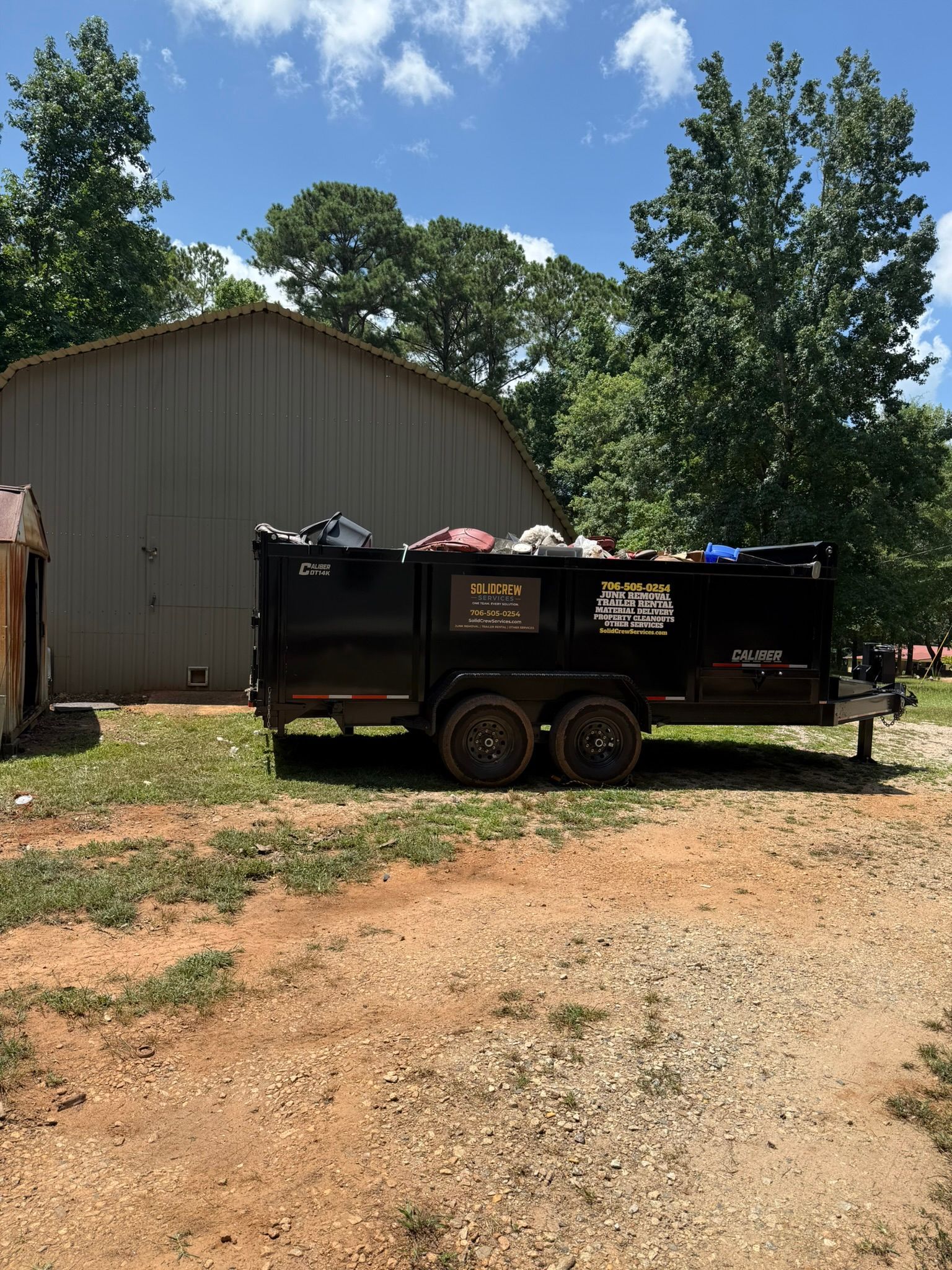Dumpster trailer filled with trash in front of a metal shed and trees on a sunny day.
