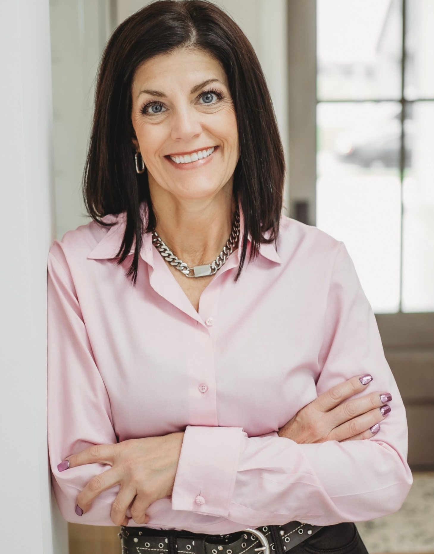 Woman with dark hair, pink shirt, silver necklace, arms crossed, smiling.