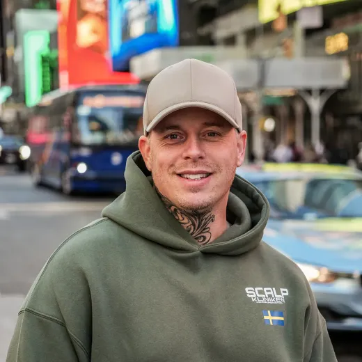 Man i beige keps och grön hoodie ler på en livlig stadsgata med suddig trafik bakom sig