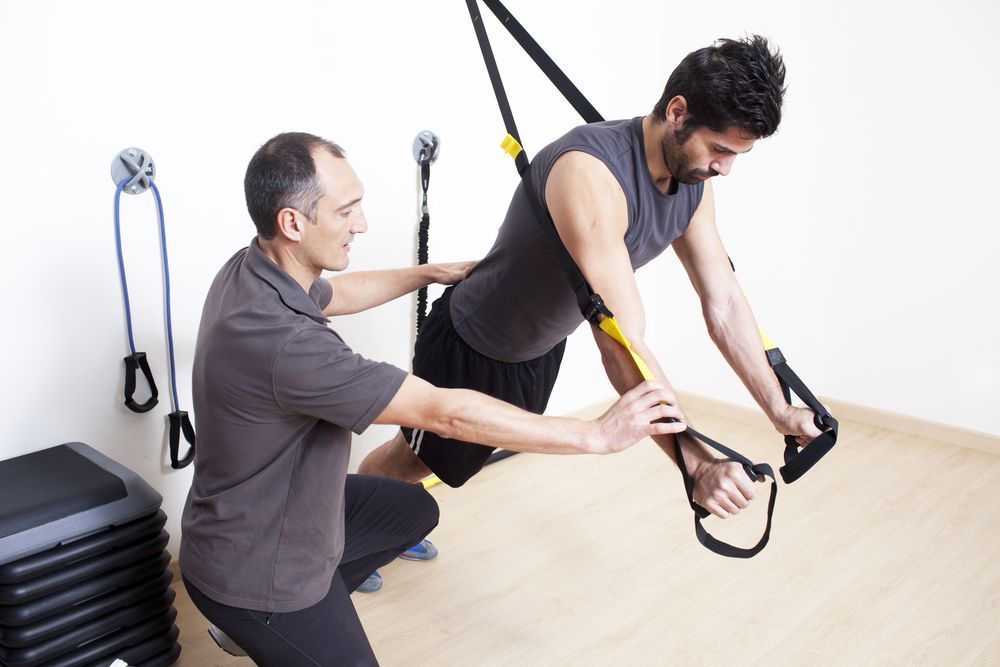 A Man Is Helping Another Man Do Trx Exercises — Caves Coastal Physiotherapy in Caves Beach, NSW