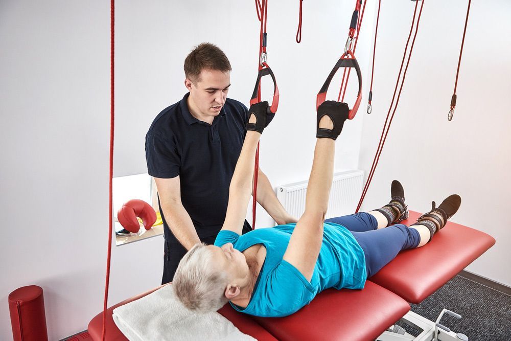 A Man Is Helping An Older Woman Do Exercises On A Bed — Caves Coastal Physiotherapy in Doyalson, NSW