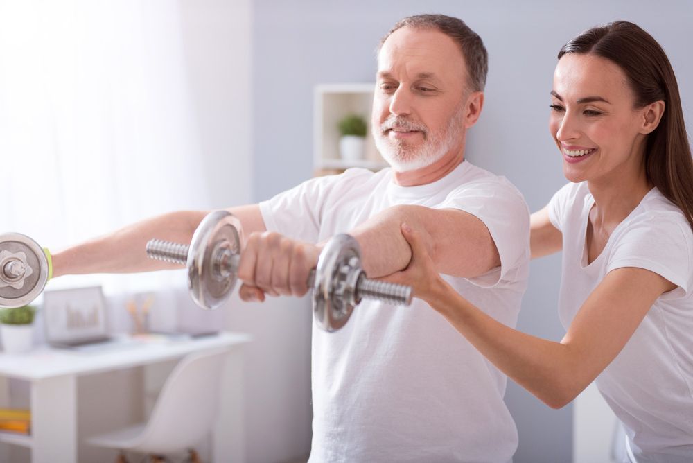 A Woman Is Helping An Older Man Lift Dumbbells — Caves Coastal Physiotherapy in Budgewoi, NSW