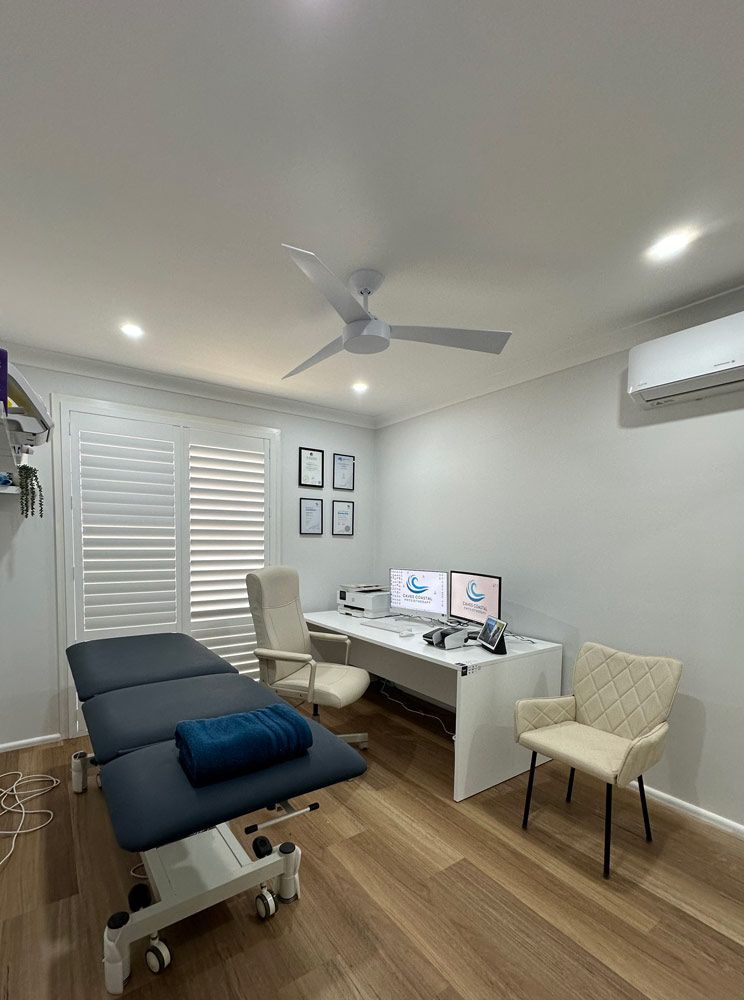 A Doctor's Office With A Bed, Desk, Chairs, Computers And A Ceiling Fan — Caves Coastal Physiotherapy in Caves Beach, NSW