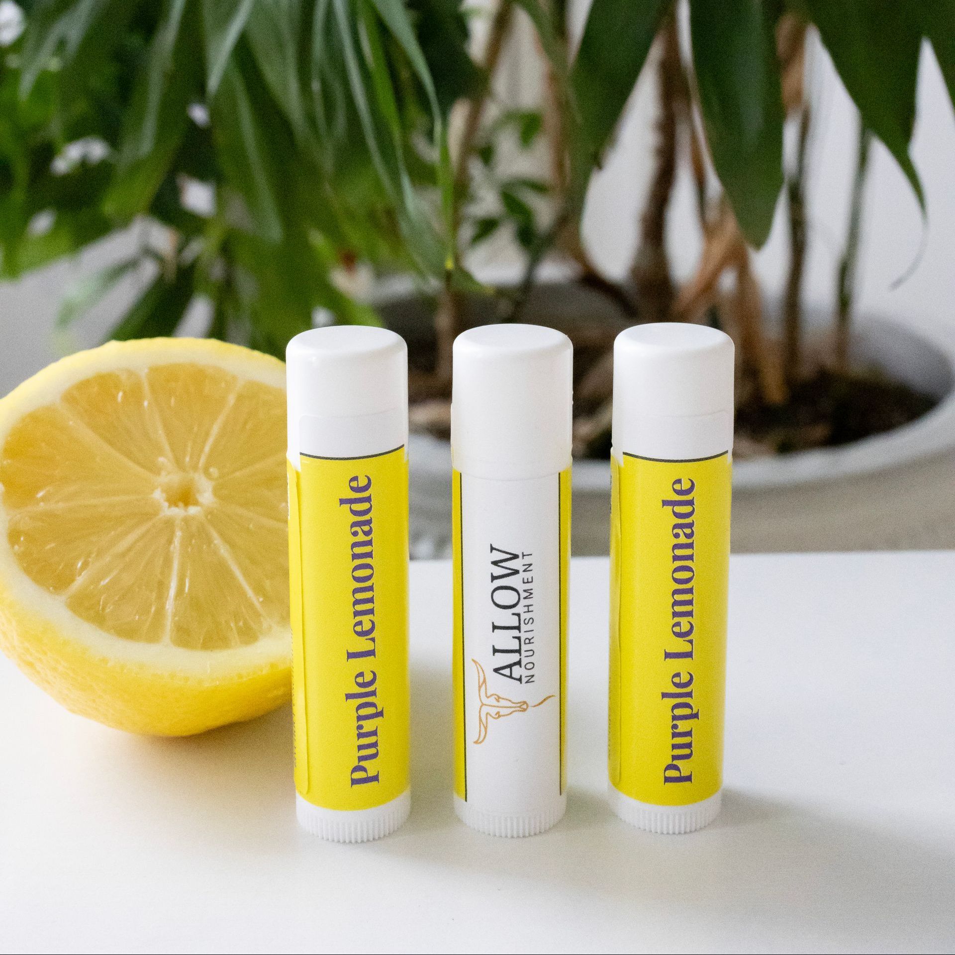 Three Allow Nourishment purple lemonade lip balms next to a slice of lemon