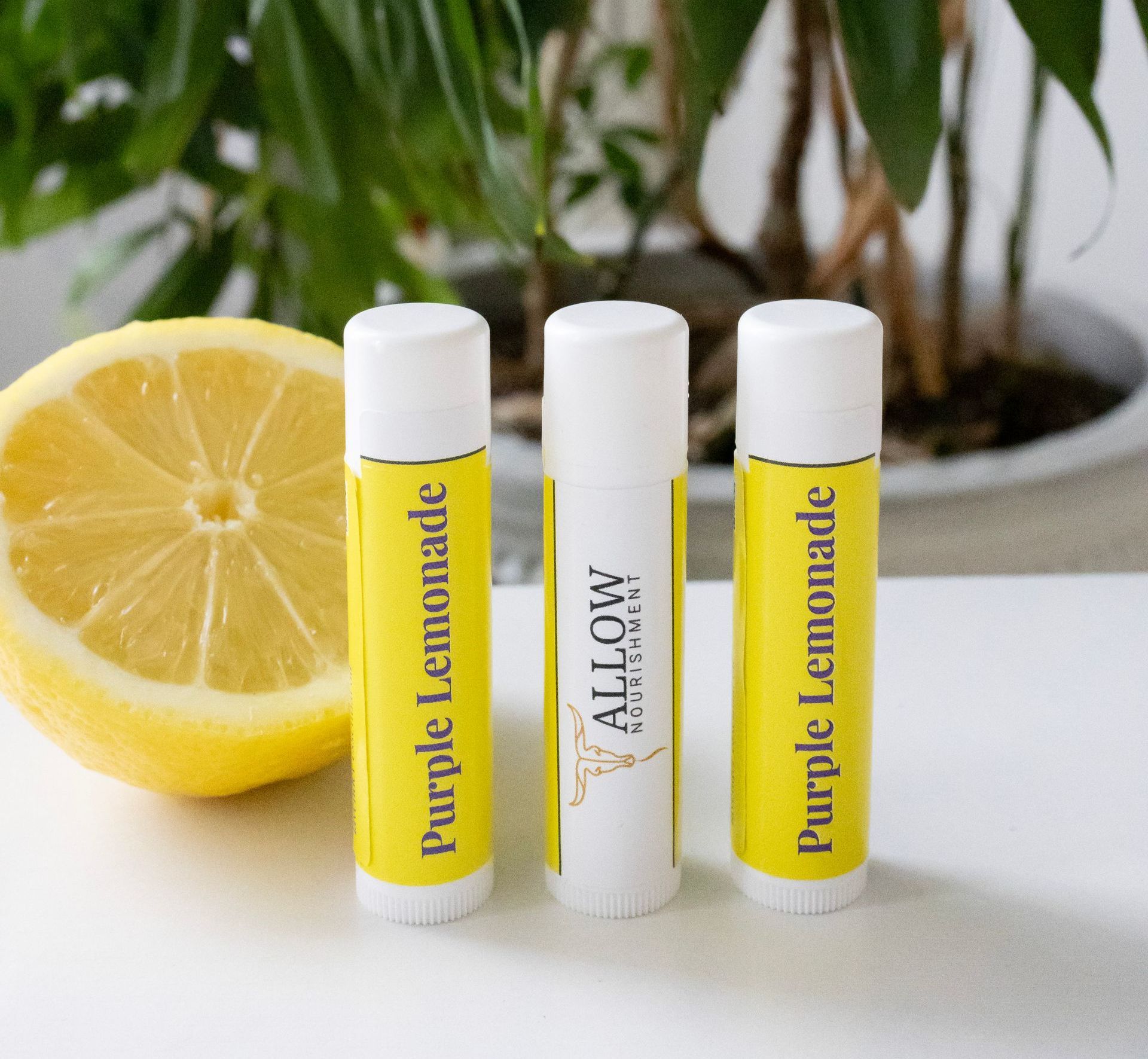 Three purple lemonade lip balms next to a slice of lemon by Allow Nourishment