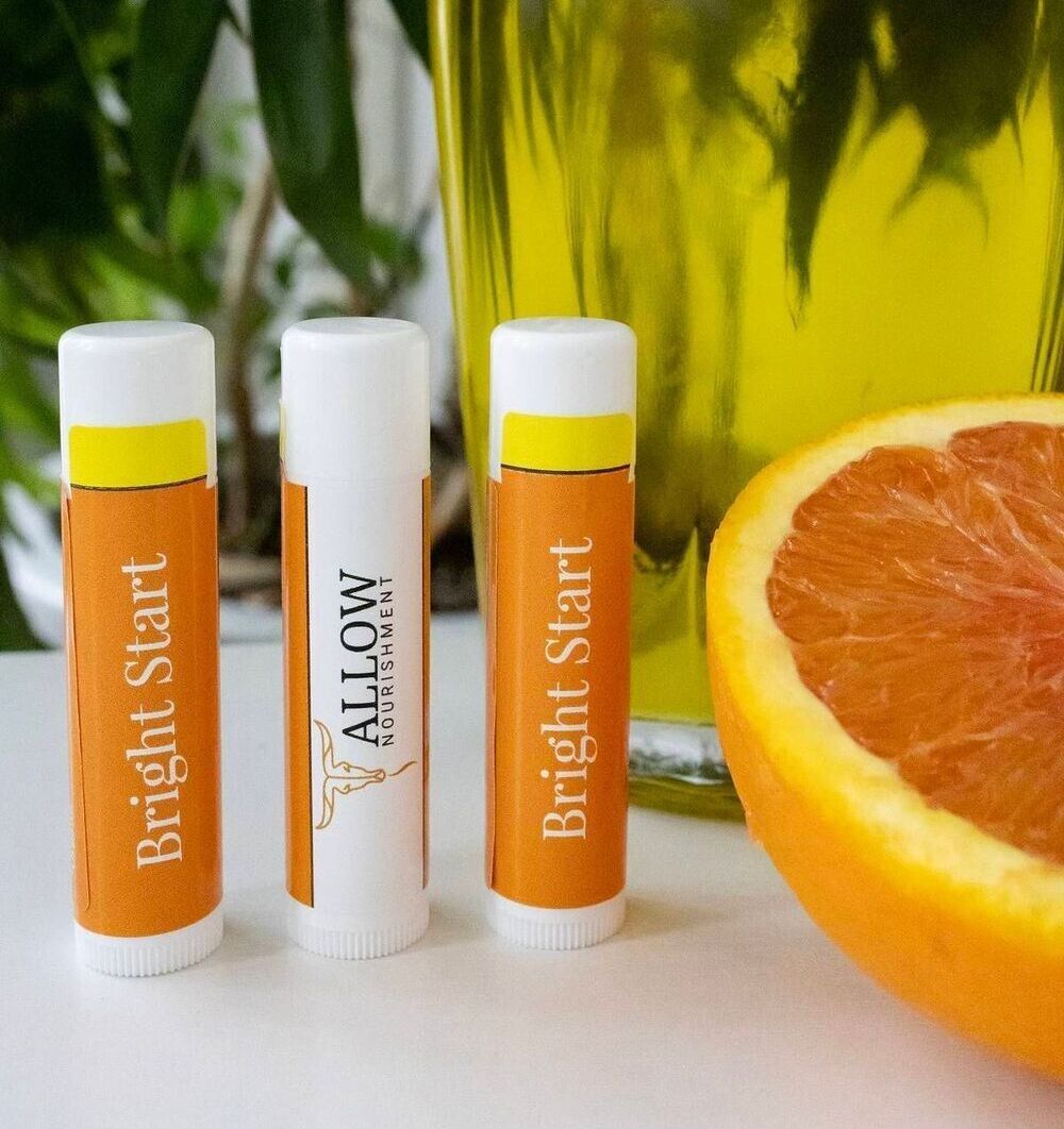 Three tubes of Allow Nourishment Bright Start Tallow lip balm next to an orange