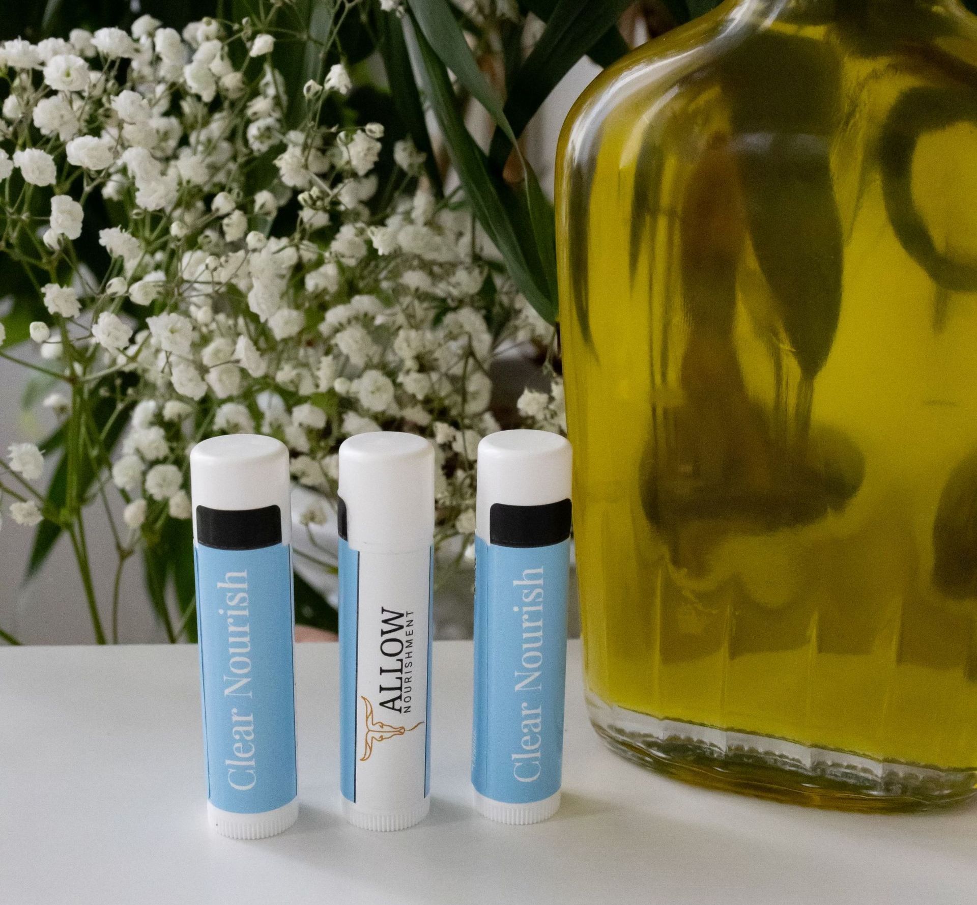 three Allow Nourish clear nourish lip balms next to a bottle of olive oil