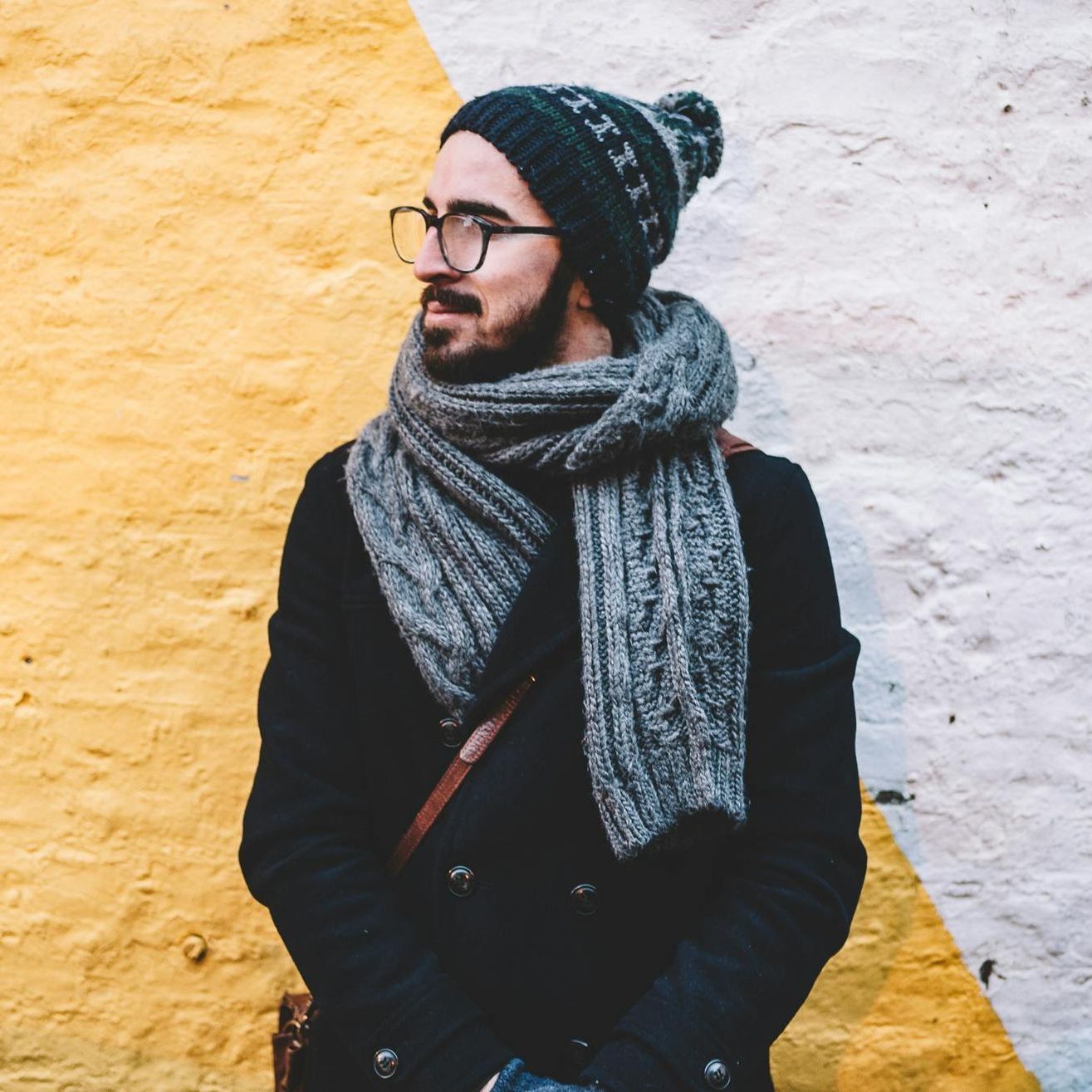 A man wearing a hat and scarf is standing in front of a yellow and white wall