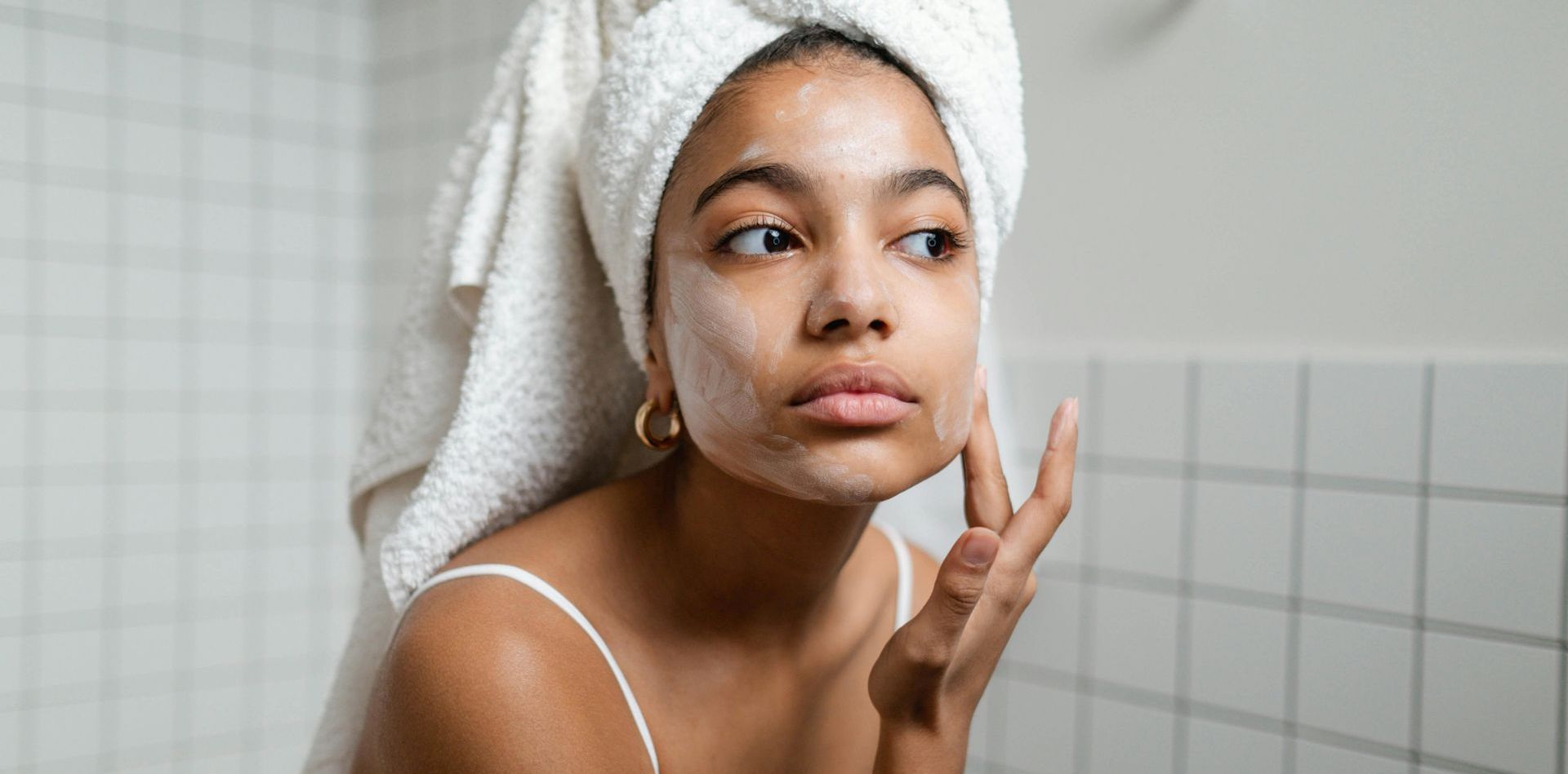 A woman with a towel wrapped around her head is looking at her face in the mirror.