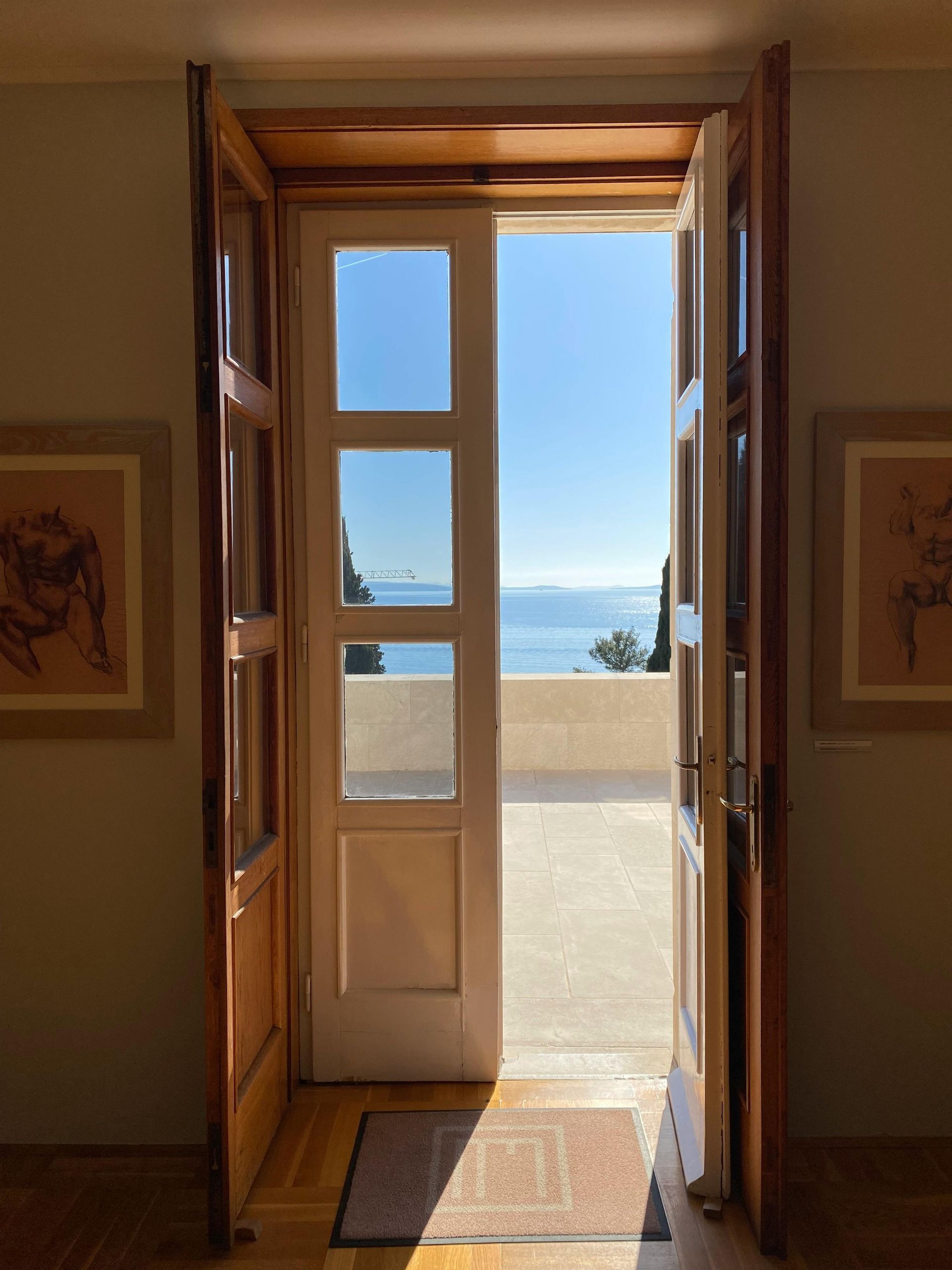 Open doorway revealing a sunny terrace with a sea view; artwork on interior walls.