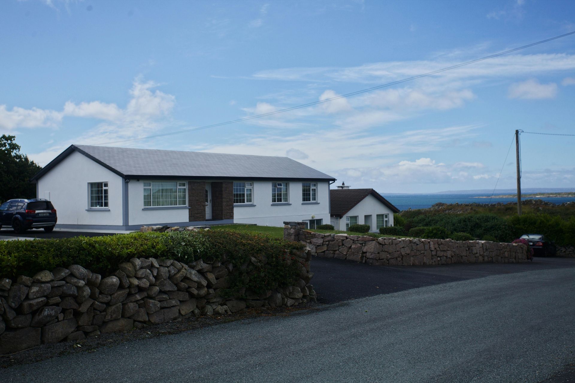 White bungalow with grey roof near a stone wall, with a view of the ocean under a blue sky.