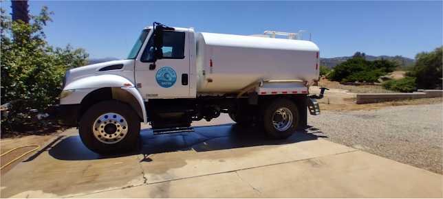 A white water truck parked on a dirt lot against a backdrop of hills, with a logo visible on the tank.