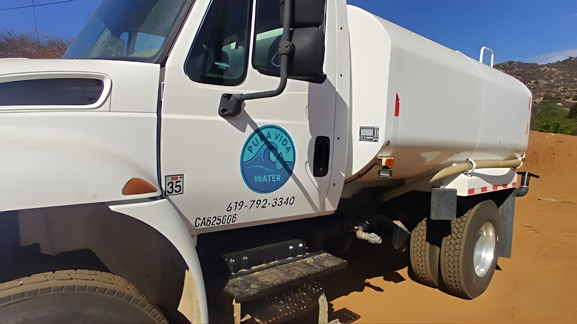 A white water tanker truck parked on a dirt lot with a hose extended towards the ground.