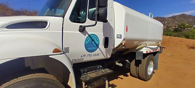 A white water truck parked on a dusty dirt road, set against a backdrop of arid, hazy mountains under a clear blue sky.