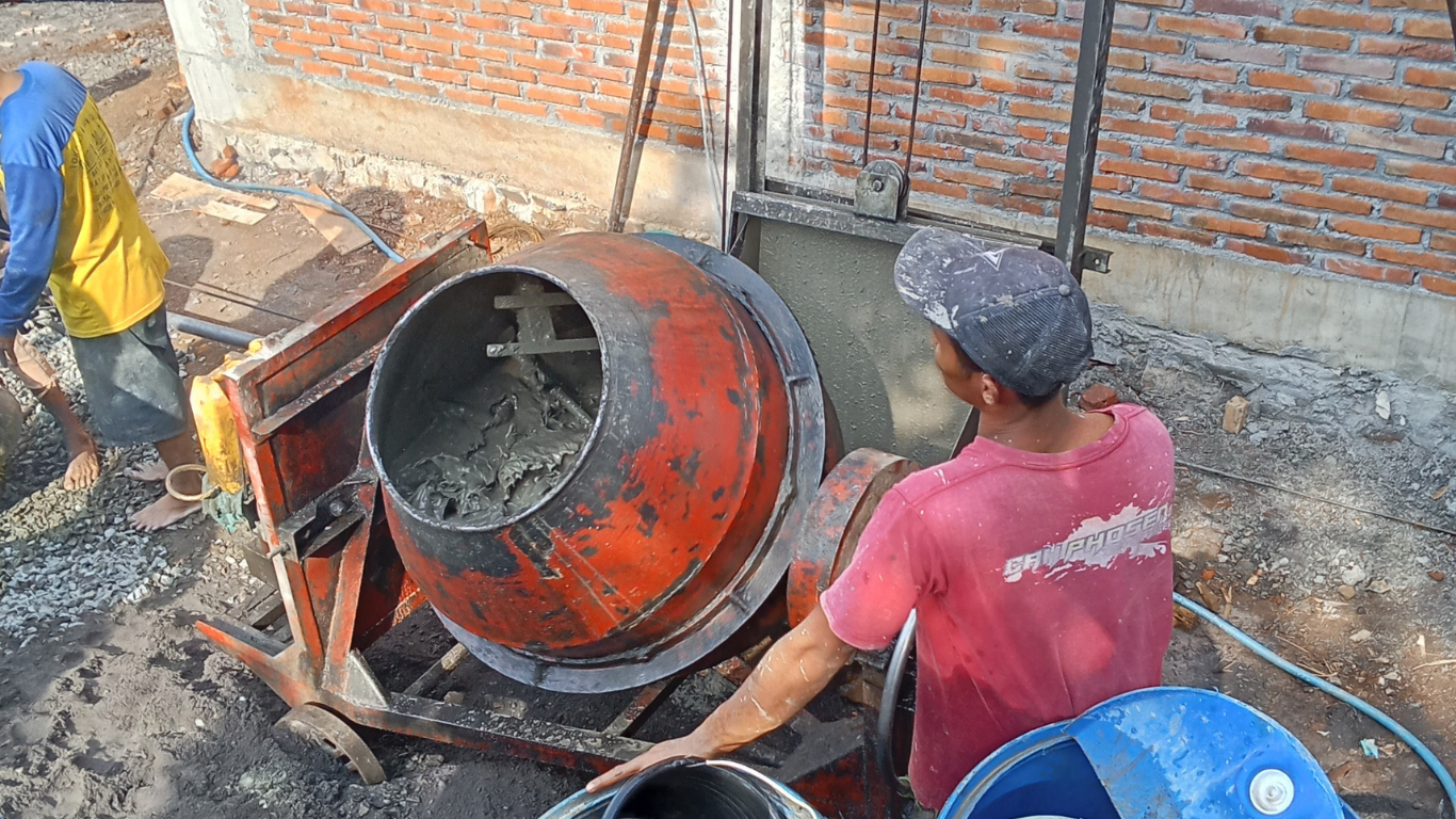 Two workers operate a portable orange cement mixer at a construction site next to a brick wall.