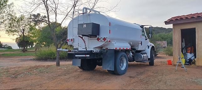 A white water tanker truck parked next to a large, metallic cylindrical water tank outdoors.