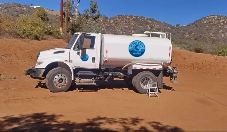 A white water truck parked on a dirt lot against a backdrop of hills, with a logo visible on the tank.