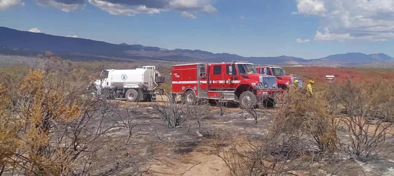 Fire trucks and a water tanker parked in a dry, scorched landscape under a blue sky.