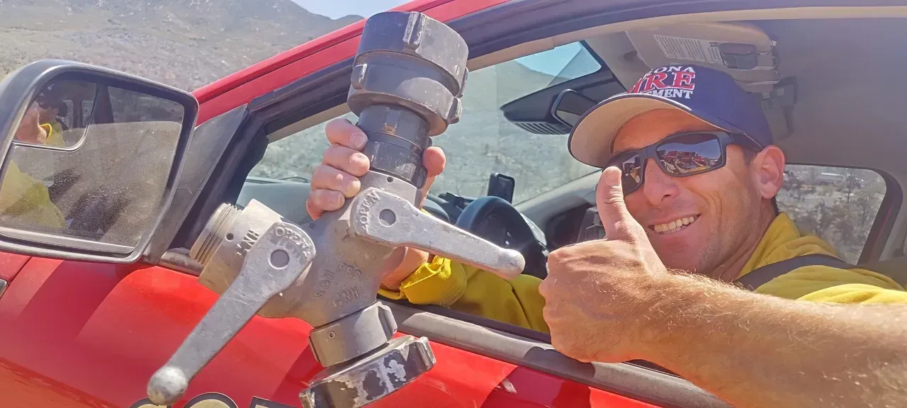 A smiling person wearing sunglasses and a cap gives a thumbs-up from a red vehicle, holding a metal fire hose nozzle.
