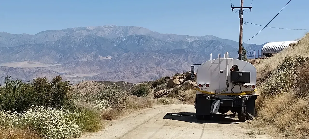 A white water truck parked on a dusty dirt road, set against a backdrop of arid, hazy mountains under a clear blue sky.