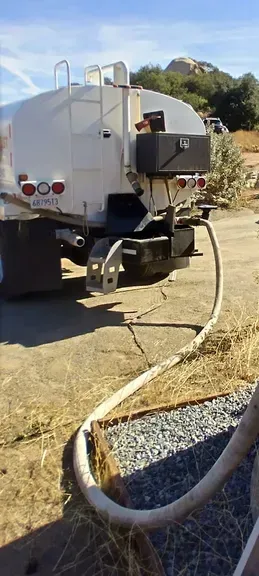 A white water tanker truck parked on a dirt lot with a hose extended towards the ground.