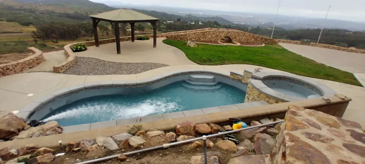 A swimming pool with an attached spa, a gazebo, and a stone retaining wall overlooking a rural landscape.