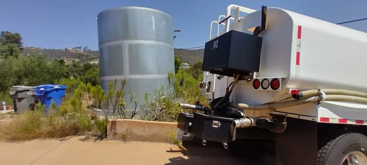 A white water truck parked next to a large, light grey industrial tank in a dusty, outdoor setting.