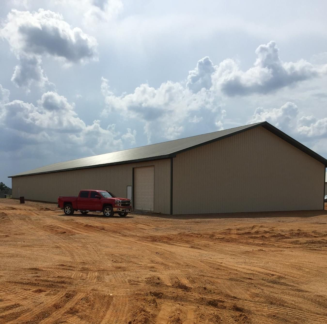 Red pickup truck parked in front of a large tan metal building, under a cloudy sky.