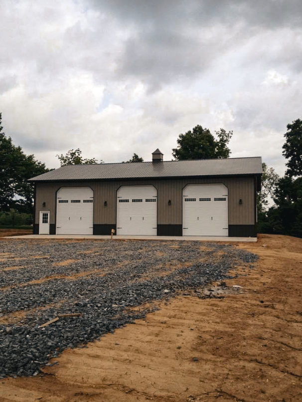 White metal building with burgundy trim and roof, gravel driveway, and a small tree.