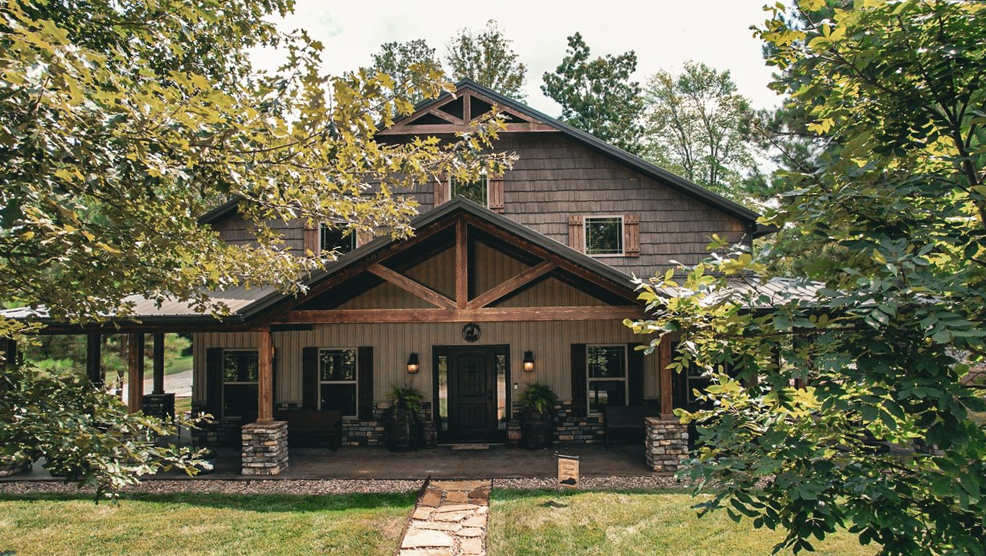 Rustic two-story cabin with a porch, stone columns, and wooden beams, nestled among trees.