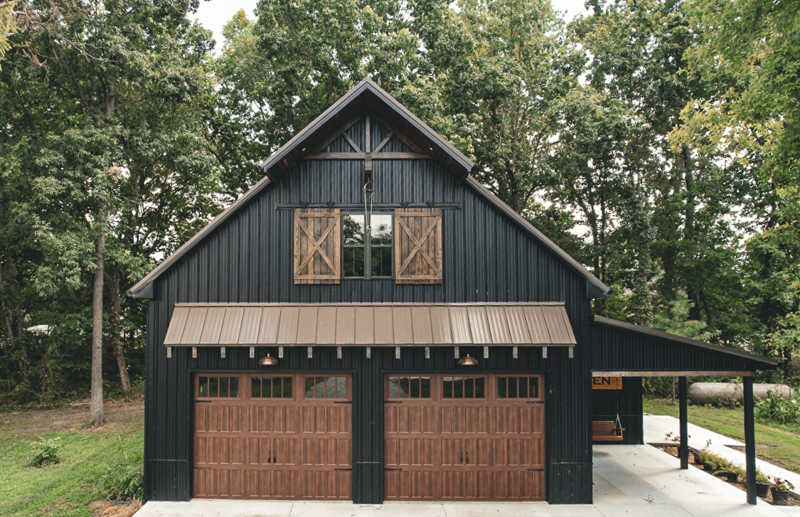 Black barn-style garage with brown doors, shutters, and awning. Lush trees in the background.