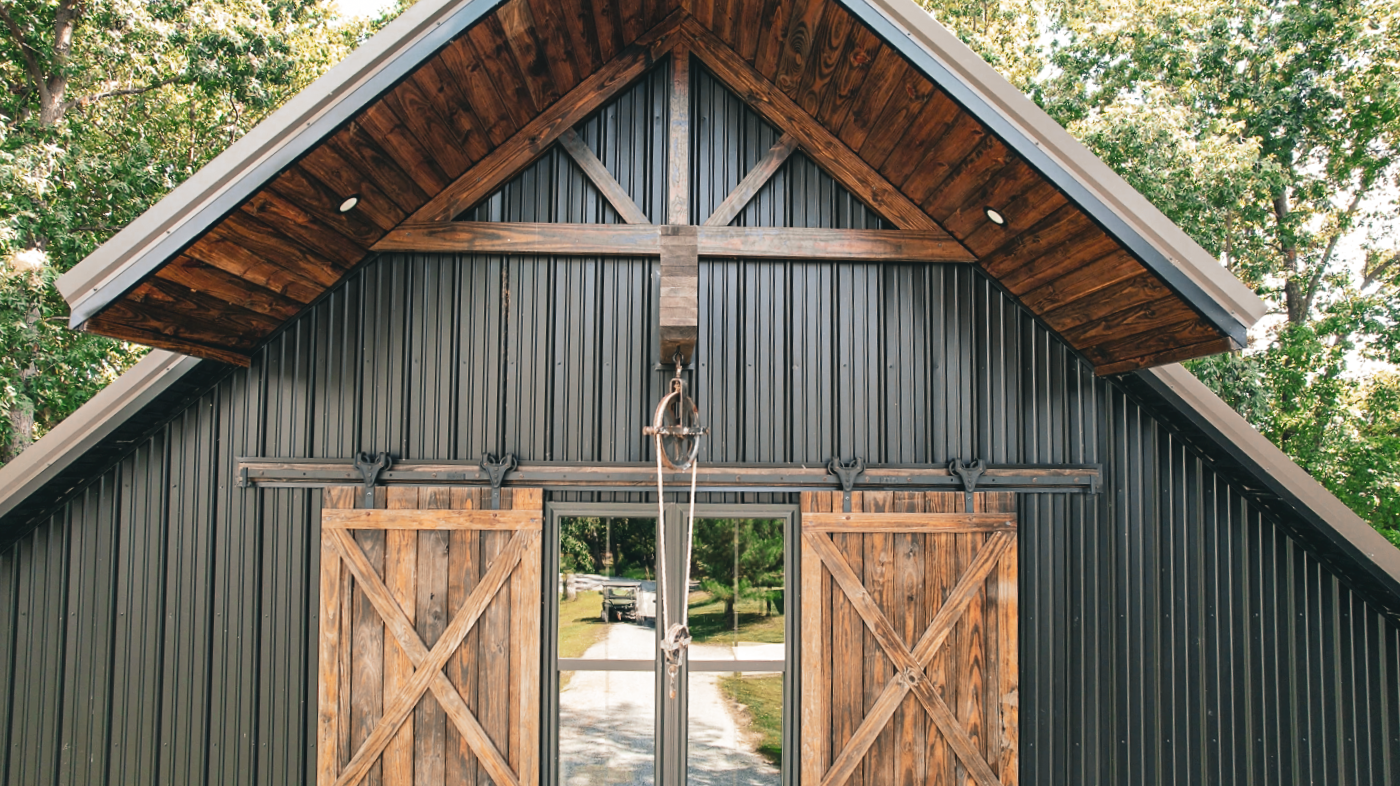 Barn-style building with a porch, two garage doors, and an adjacent small shed under a blue sky.