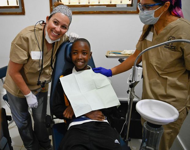 A young boy is sitting in a dental chair with two female dentists standing around him