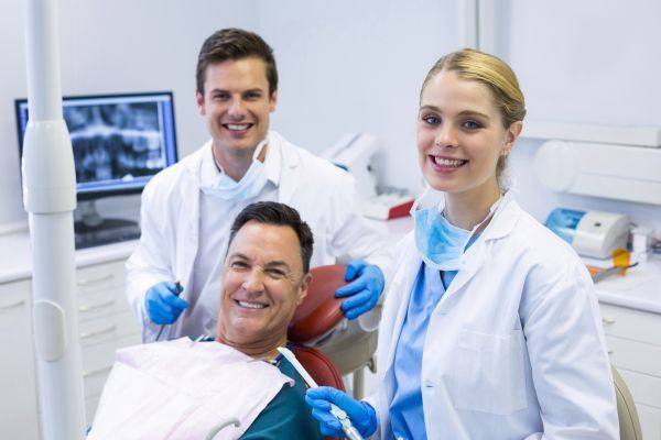 Man in dentist chair smiles, two dental professionals smile behind him in a dental office.