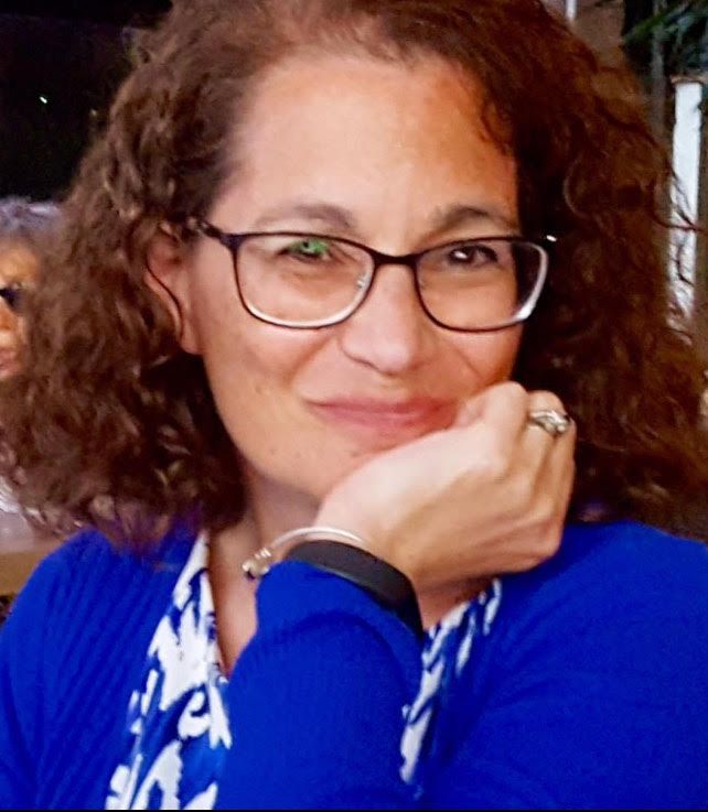 Woman wearing glasses, a purple sweater, and a necklace, smiles at the camera.