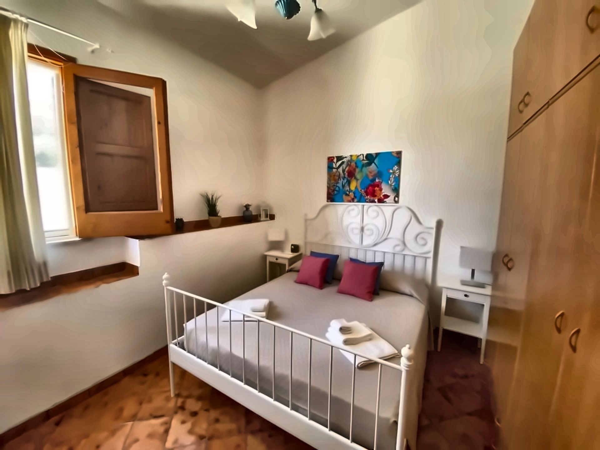 Camera da letto con letto bianco, finestra e armadio in legno. Cuscini blu e rossi.