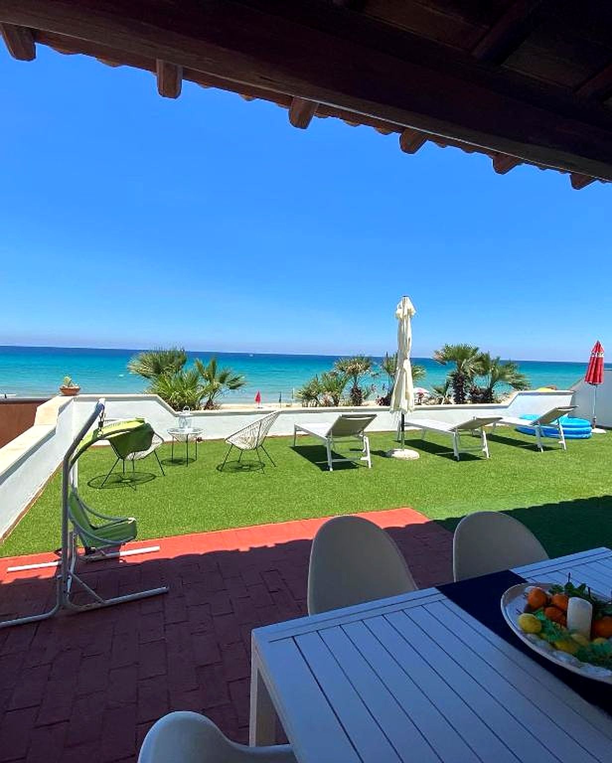Patio con vista sulla spiaggia e sul cielo azzurro. Sedie da giardino, altalena, tavolo apparecchiato con frutta.